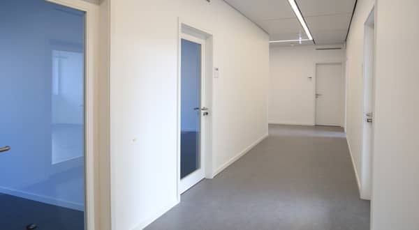 Vue d'ensemble d'un couloir d'entreprise, trois portes intérieures vitrées, une porte pleine en bois couleur blanche dans le fond.