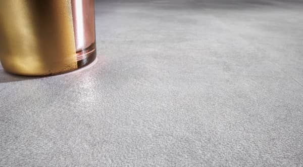 Zoom sur un sol en PVC gris moucheté, sous forme de dalles, bouteille décorative dorée posée dessus.