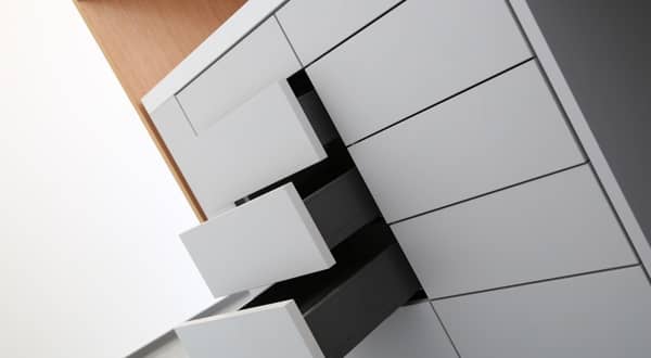 Photo prise de biais d'un caisson de rangement gris clair avec 2 colonnes de 5 tiroirs, sans poignées, sous un bureau en bois.