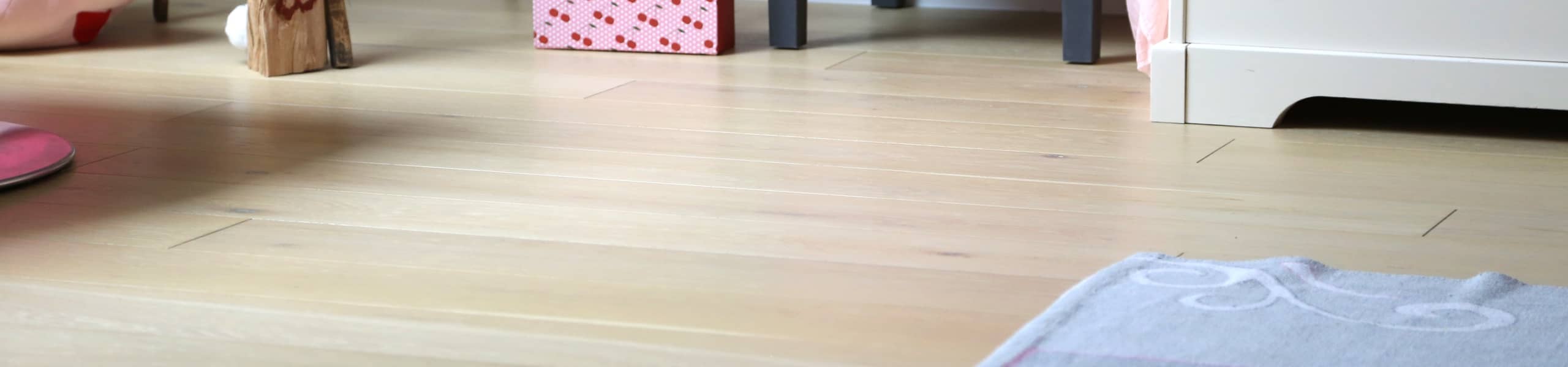 Vue d'ensemble d'une chambre de petite fille, mise en avant d'un parquet massif couleur clair et vitrifié, tapis, jouets, lit.