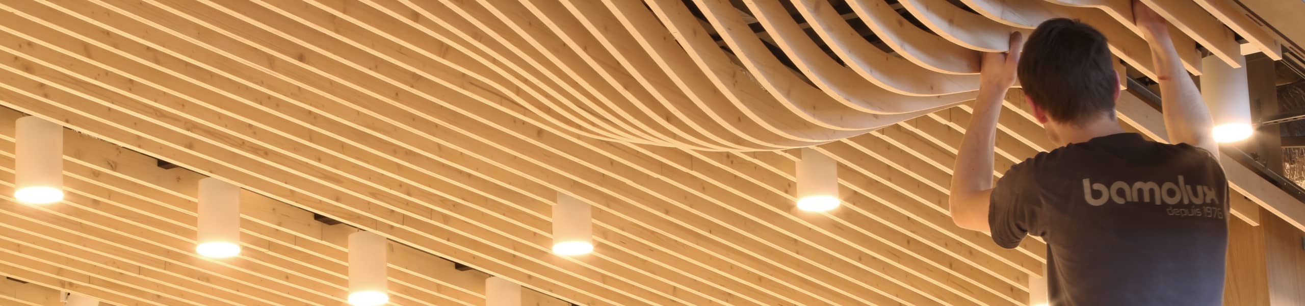 Poseur Bamolux qui installe un plafond suspendu en lames de bois clair avec formes de bulle à certain endroit et spots intégrés.